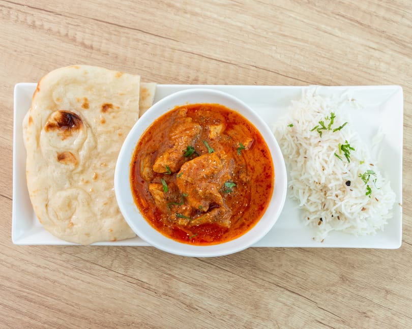 Chicken curry with naan bread and basmati rice, garnished with cilantro, on a wooden table.