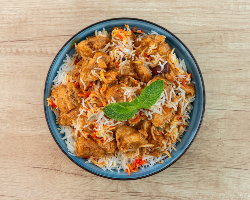Chicken biryani with rice, chicken pieces, and mint leaves in a blue bowl on a wooden table.