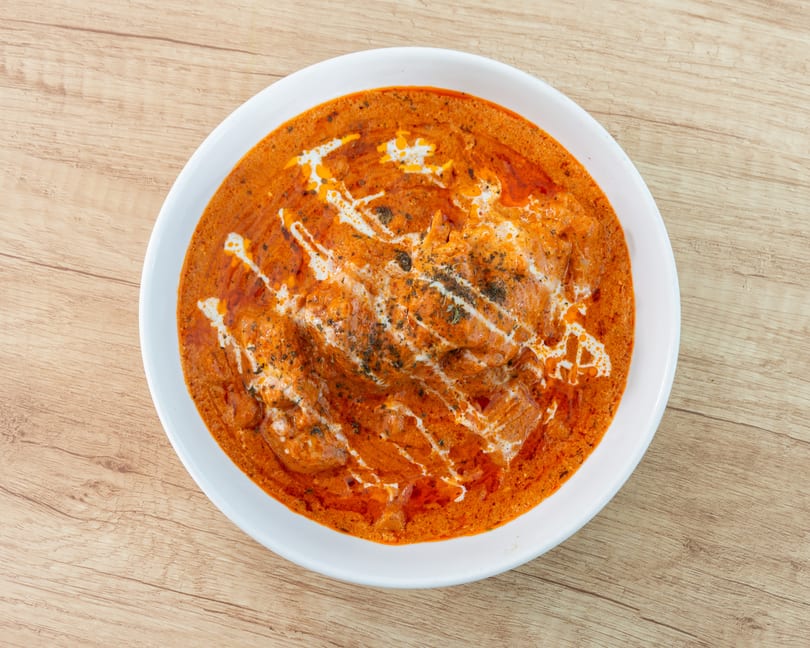 Overhead view of a white bowl filled with orange butter chicken, drizzled with cream and herbs.