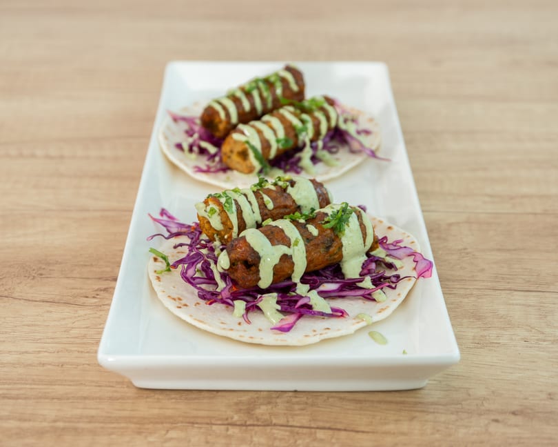 Three kebabs on tortillas with red cabbage and green sauce, served on a white rectangular plate.