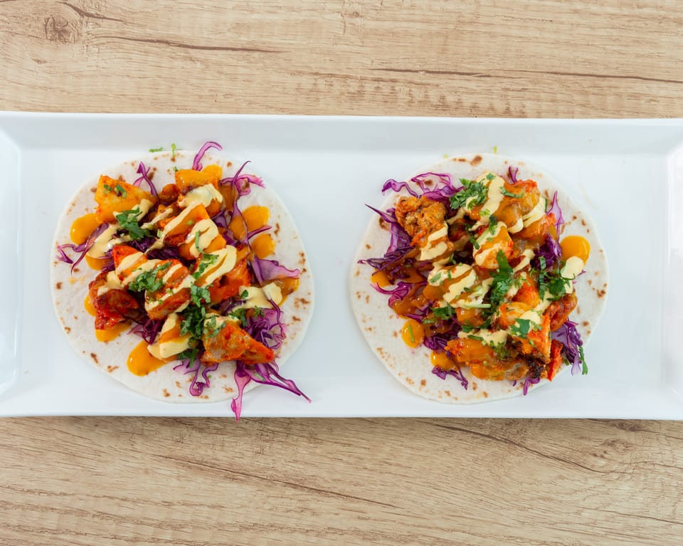 Two vibrant tacos with orange chicken, purple cabbage, and creamy sauces on a white platter.
