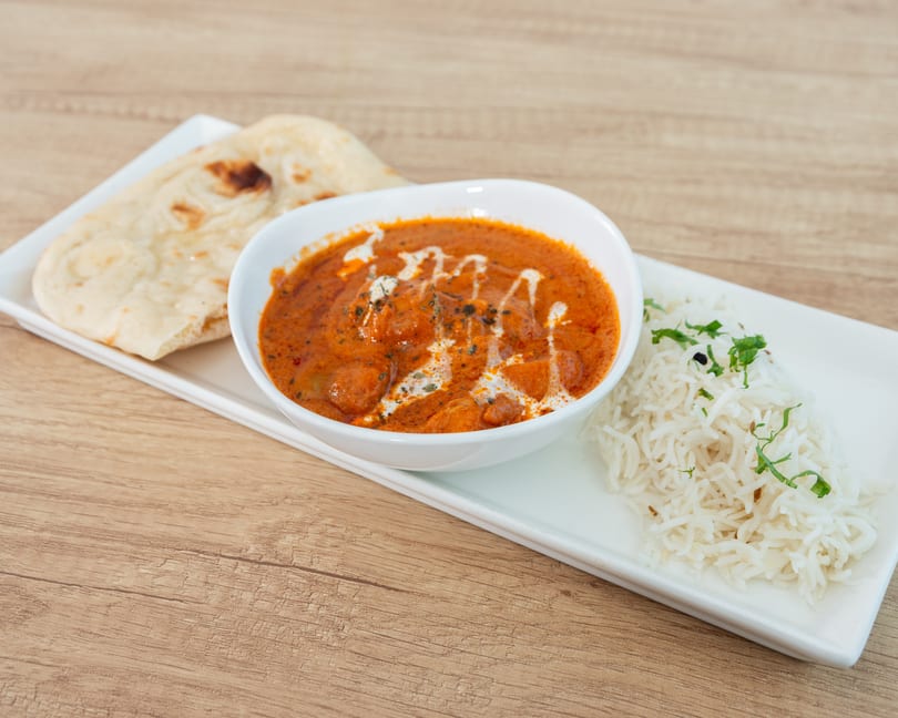 A bowl of butter chicken with naan bread and rice on a white rectangular plate.