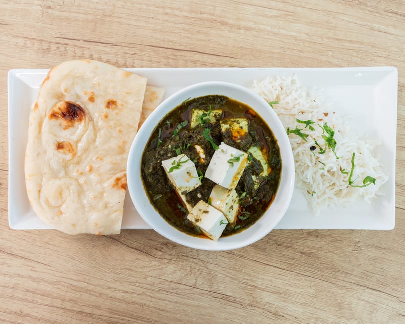 Palak Paneer with rice and naan bread on a white rectangular plate on a wooden table.